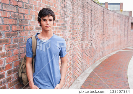 Man in 20s leaning against red brick wall beside campus walkway carrying khaki backpack, copy space 135307584