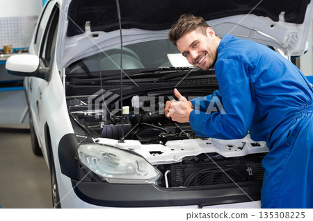 Male mechanic in garage leaning into car engine under open hood showing support rod, copy space 135308225
