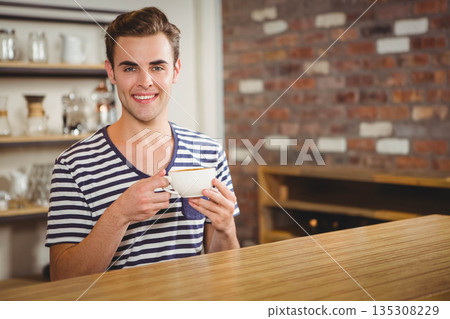 Mid adult man holding white ceramic coffee cup behind counter in cafe with exposed brick wall 135308229