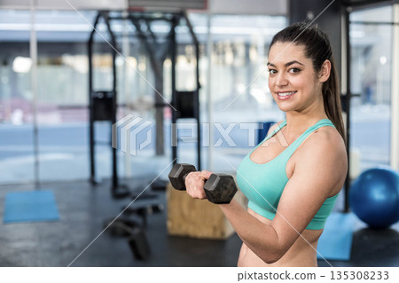 Woman in turquoise sports bra and leggings curling dumbbell at chest in studio, copy space Woman in turquoise sports bra and leggings curling dumbbell at chest in studio, copy space 135308233