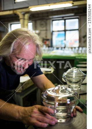 Male artisan wearing apron examining glass vessel on turntable at workshop workbench, copy space 135308244