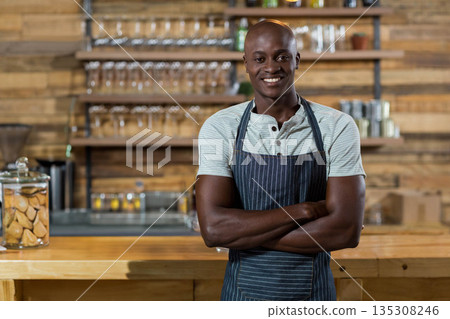 Standing African American man wearing apron behind bar counter in cafe with glass jar cookies Standing African American man wearing apron behind bar counter in cafe with glass jar cookies 135308246