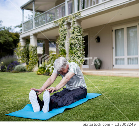 Senior male performing forward stretch on blue exercise mat on backyard lawn wearing sneakers 135308249