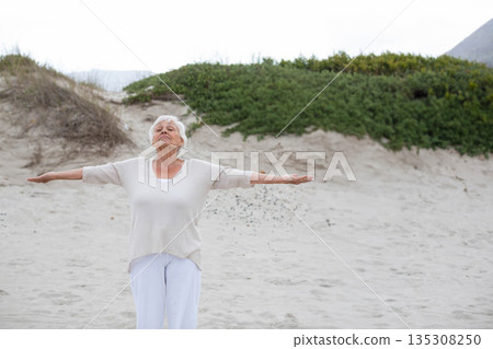 Senior woman stretching arms sideways and taking deep breath on sandy beach with dune grass Senior woman stretching arms sideways and taking deep breath on sandy beach with dune grass 135308250