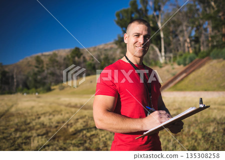 Man wearing red T-shirt, lanyard writing notes with pen on clipboard in meadow, copy space Man wearing red T-shirt, lanyard writing notes with pen on clipboard in meadow, copy space 135308258