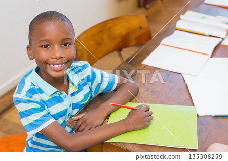 African American boy student sitting at desk in classroom holding red pencil over green notebook African American boy student sitting at desk in classroom holding red pencil over green notebook 135308259