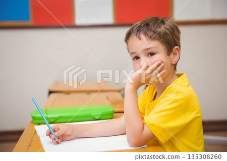 Boy sitting at school desk wearing yellow t-shirt and holding blue pencil over paper, copy space 135308260