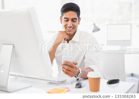 Indian man wearing white shirt leaning forward at office desk, checking smartphone near monitor Indian man wearing white shirt leaning forward at office desk, checking smartphone near monitor 135308263