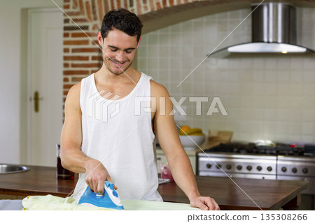 Man ironing shirt with steam iron on wooden kitchen island beside metal stove and tiled backsplash 135308266