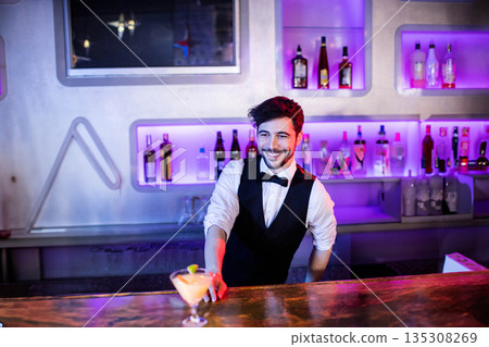 Male bartender in black vest smiling and standing behind bar counter with frosted cocktail glass Male bartender in black vest smiling and standing behind bar counter with frosted cocktail glass 135308269