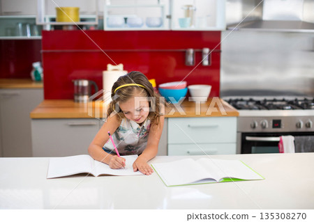School-age girl writing in notebook at kitchen island with colorful mixing bowls, metal kettle 135308270