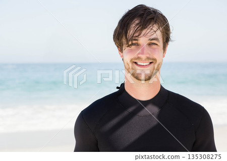 Man standing near waterline on sandy beach wearing black wetsuit and smiling, copy space Man standing near waterline on sandy beach wearing black wetsuit and smiling, copy space 135308275