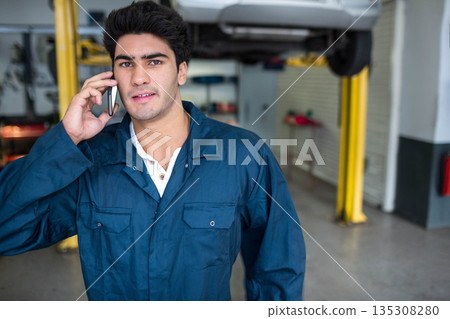 Indian male mechanic standing at vehicle repair garage talking on smartphone beside hydraulic lift Indian male mechanic standing at vehicle repair garage talking on smartphone beside hydraulic lift 135308280
