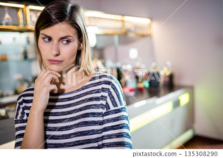 Woman leaning on counter resting chin on hand while gazing right at bar glassware bottles 135308290