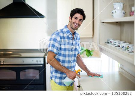 Man cleaning white countertop with green cloth and spray bottle in home kitchen near stove 135308292