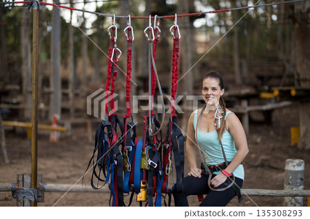 Female participant kneeling wearing harness next to carabiners on wooded ropes course, copy space Female participant kneeling wearing harness next to carabiners on wooded ropes course, copy space 135308293