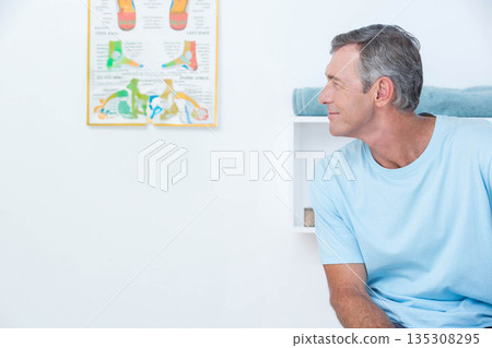 Mature man sitting on cushioned table in medical office viewing foot anatomy chart, copy space Mature man sitting on cushioned table in medical office viewing foot anatomy chart, copy space 135308295
