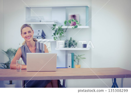 Woman smiling while sitting at wooden desk with laptop, water glass, plants, binders and folders 135308299