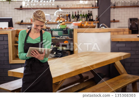 Female barista wearing black apron tapping tablet near communal table in coffee shop, copy space Female barista wearing black apron tapping tablet near communal table in coffee shop, copy space 135308303