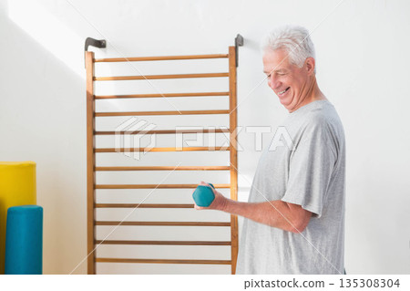 Senior man lifting teal hand weight beside exercise ladder with foam rollers in gym, copy space Senior man lifting teal hand weight beside exercise ladder with foam rollers in gym, copy space 135308304