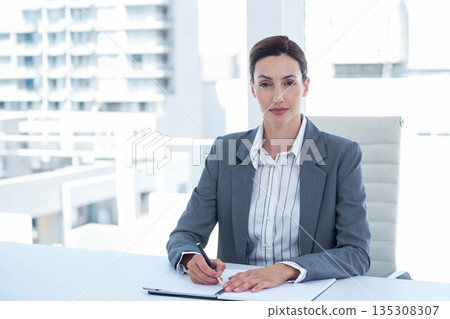 Middle-aged professional woman writing on notepad with black pen at desk near balcony railing Middle-aged professional woman writing on notepad with black pen at desk near balcony railing 135308307