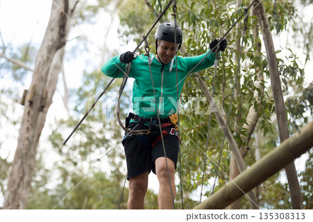 Man balancing on suspended wooden beam in forest, gripping steel cables with helmet and harness 135308313