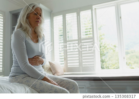 Senior woman clutching abdomen sitting on bed in bedroom with shutters and pillows, copy space Senior woman clutching abdomen sitting on bed in bedroom with shutters and pillows, copy space 135308317