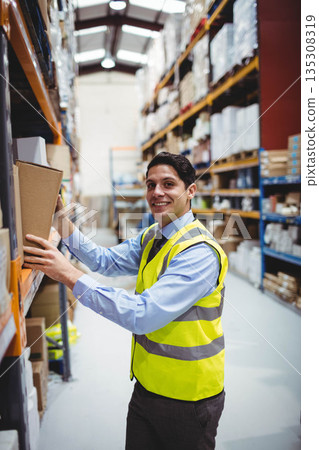 Male worker wearing safety vest reaching to retrieve cardboard box from shelving in storage aisle Male worker wearing safety vest reaching to retrieve cardboard box from shelving in storage aisle 135308319