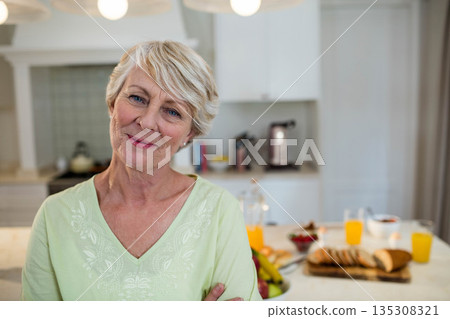 Senior woman standing at kitchen island preparing breakfast with orange juice, toaster and fruit 135308321