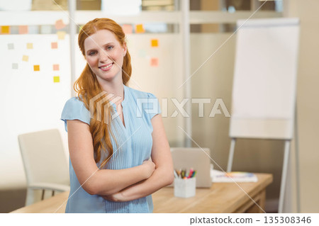 Red-haired woman crossing arms at desk in office with sticky notes and flipchart 135308346