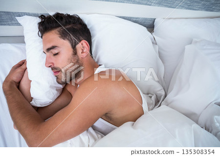 Man in his 20s sleeping in bedroom on bed wearing tank top hugging pillow and comforter 135308354