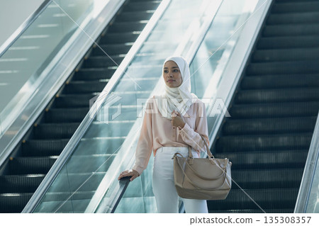 Woman in twenties wearing white hijab descending glass escalator in hub holding beige tote bag 135308357