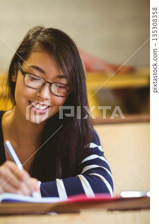 Asian woman sitting at desk in study room wearing glasses writing in notebook with silver pen 135308358