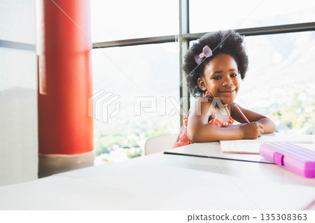 African American girl writing in notebook at table by window with pink pencil case, copy space African American girl writing in notebook at table by window with pink pencil case, copy space 135308363