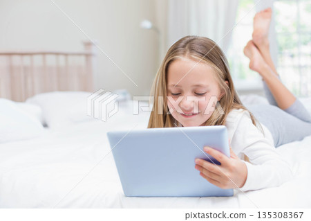 Schoolgirl lying on bed holding tablet under sheer curtains beside lamp in bedroom 135308367