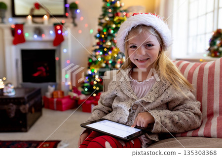Girl wearing Santa hat using tablet at home near Christmas tree, gifts and fireplace, copy space 135308368