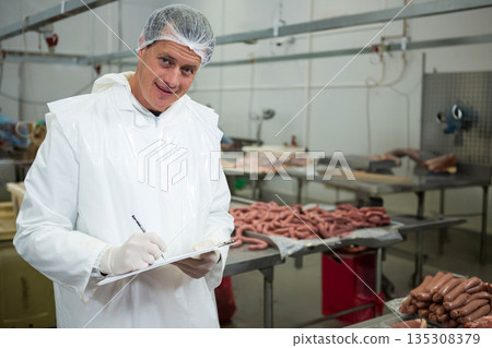 Male inspector wearing coat examining sausage trays at meat plant holding clipboard, copy space 135308379