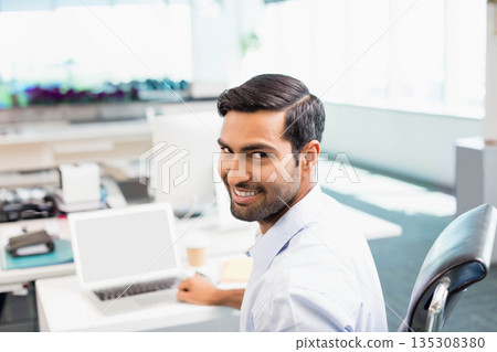 Asian man turning in swivel chair smiling at laptop on white desk in open-plan office 135308380