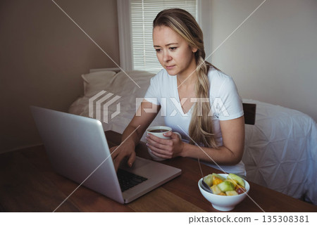 Mid adult woman wearing white T-shirt working on silver laptop on bedroom table holding ceramic mug 135308381