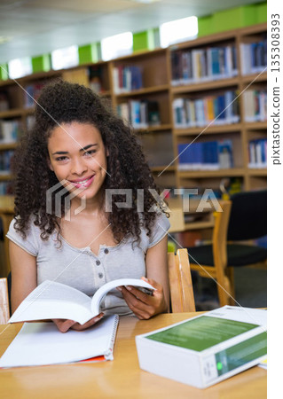 African American woman reading paperback and jotting notes in notebook by textbook on library table 135308393