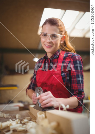 Woman wearing safety glasses and red apron using hand plane on wood block at workshop bench 135308404