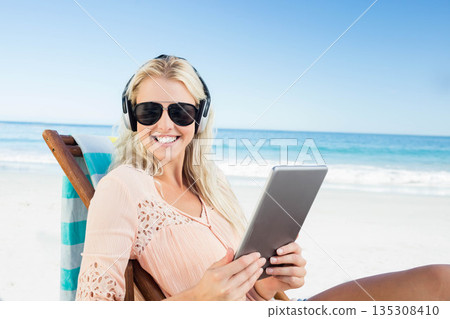 Blonde woman resting on towel-draped lounger using tablet wearing sunglasses, headphones on beach 135308410