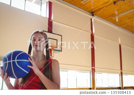 Teenage girl wearing red sports top holding basketball by hoop in gymnasium with blinds, copy space Teenage girl wearing red sports top holding basketball by hoop in gymnasium with blinds, copy space 135308413