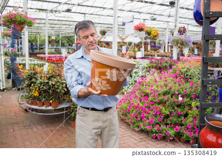 Man inspecting terracotta planter inside greenhouse near potted flowering plants 135308419