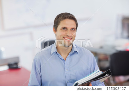 Man wearing light blue shirt standing in bright office space holding black ring binder and papers Man wearing light blue shirt standing in bright office space holding black ring binder and papers 135308427