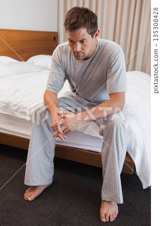Man wearing gray pajamas sitting on bed edge against wooden headboard in bedroom, gazing downward 135308428