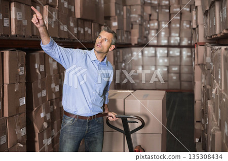 Male warehouse worker in storage aisle gripping pallet jack, pointing at barcode boxes on shelves 135308434
