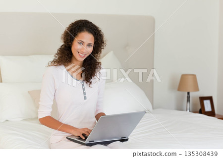 Woman sitting on bed with white linens wearing loungewear holding silver laptop at home, copy space Woman sitting on bed with white linens wearing loungewear holding silver laptop at home, copy space 135308439
