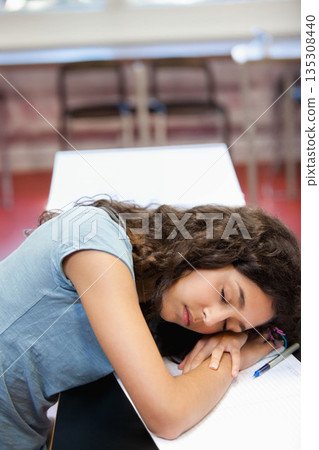 Female student resting head on arms atop notebook at classroom desk amid chairs with blue pen 135308440