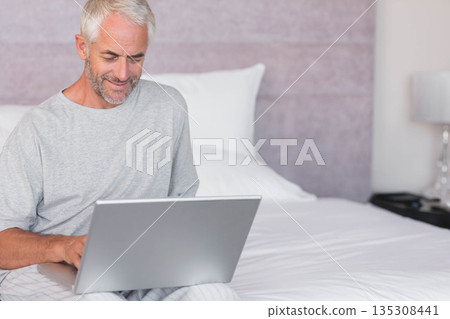 Middle aged man sitting cross-legged on bed at home using laptop with metallic lamp and tray 135308441